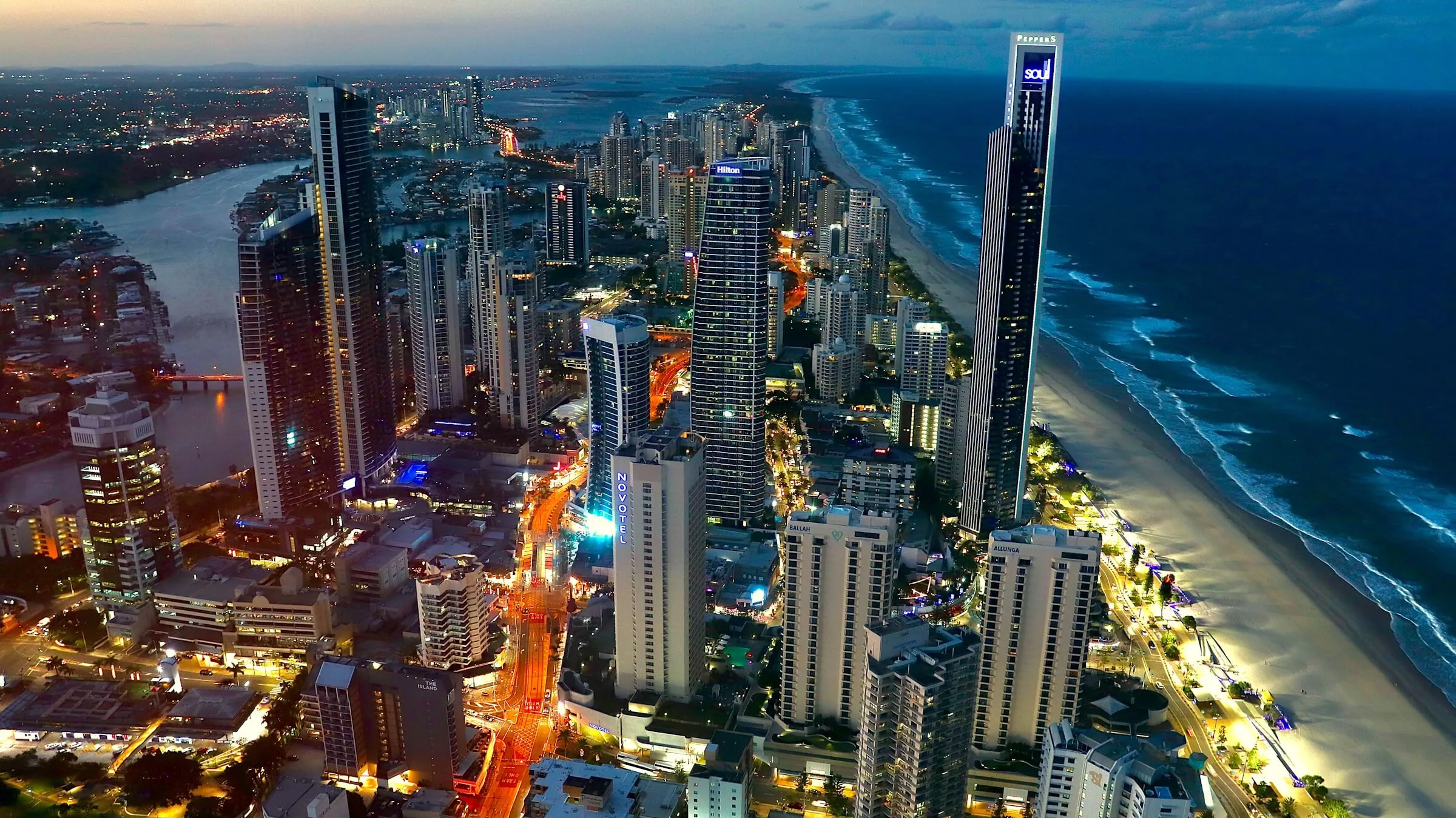 Surfers Paradise from high above at dusk: lit towers between the Nerang River on the left and the beach and ocean on the right